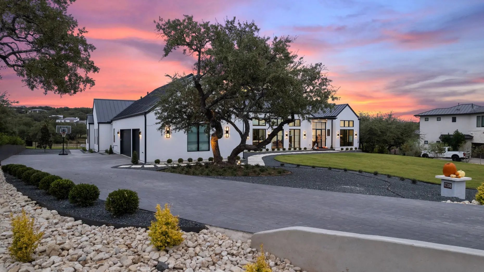White farmhouse with a dark metal roof at sunset, behind a live oak and a forecourt of gravel and boulders.