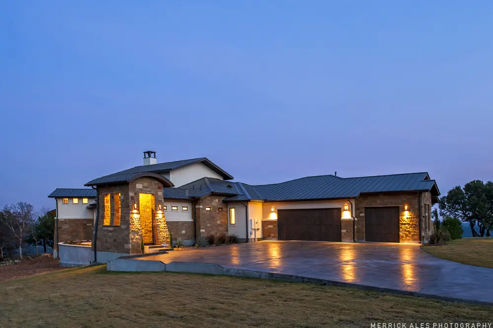 Front elevation at dusk of a contemporary stone and stucco house with a metal roof and lit garage bays across a wide drive.