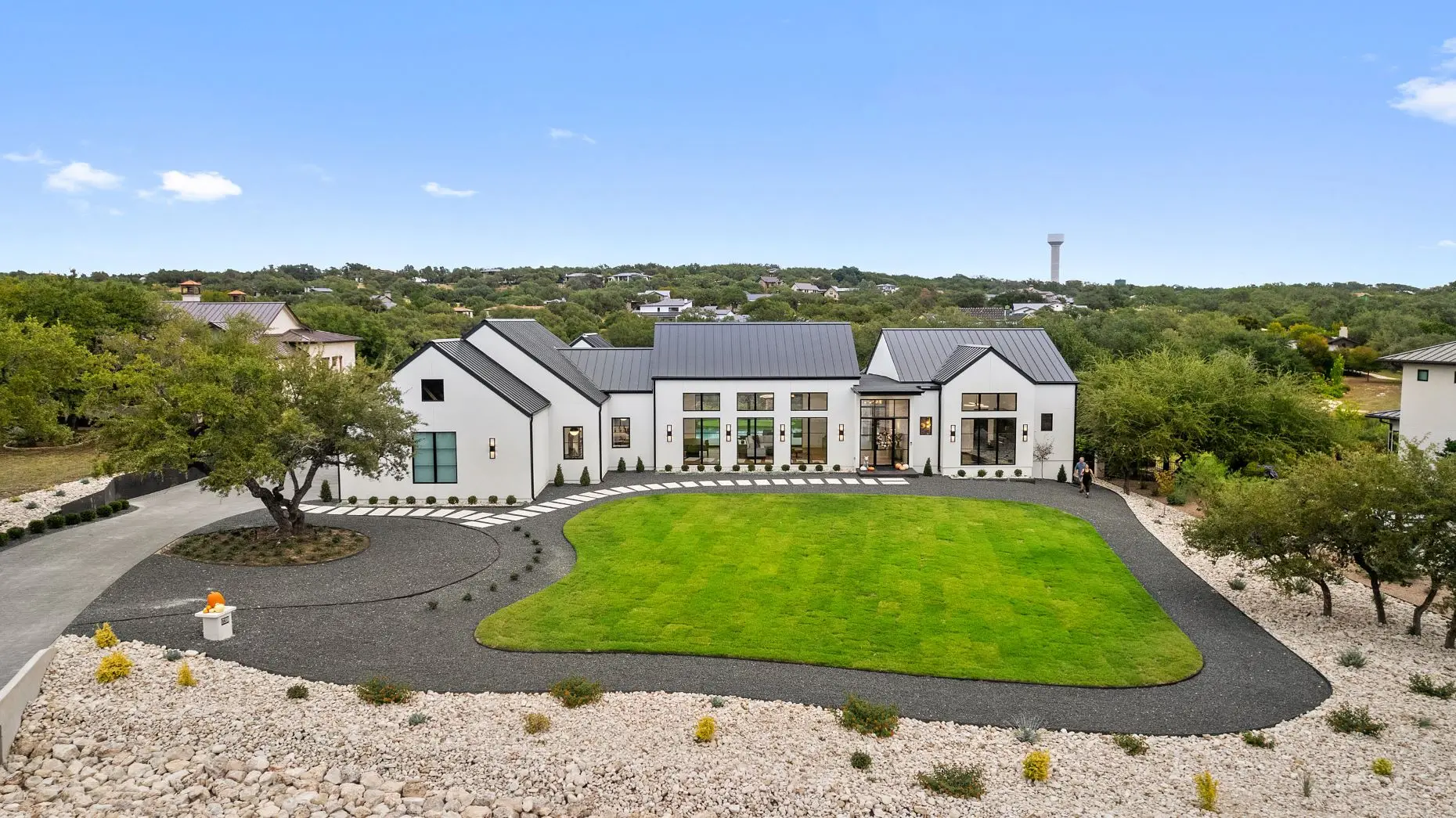 White farmhouse with a dark metal roof at sunset, behind a live oak and a forecourt of gravel and boulders.