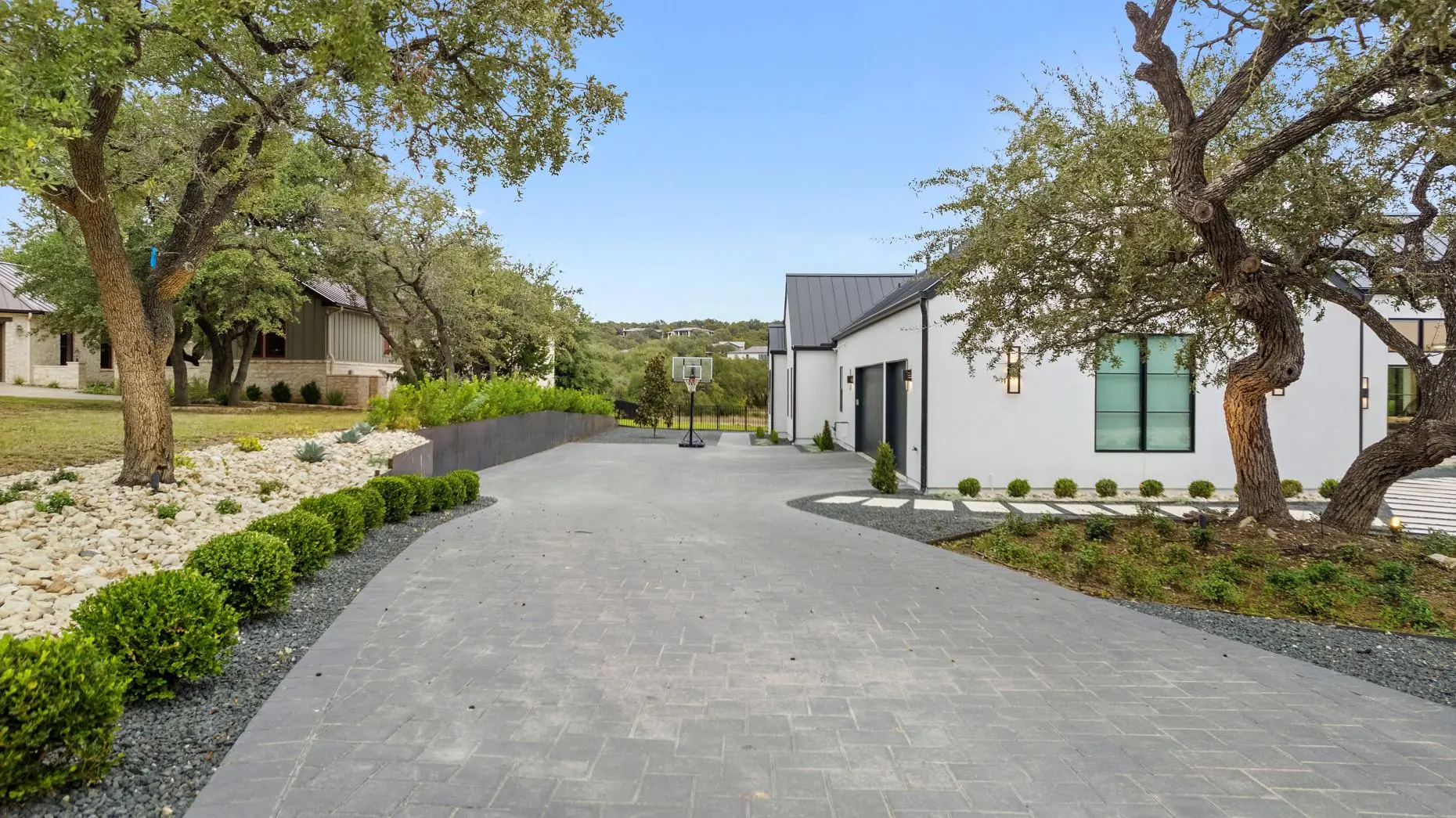 White farmhouse with a dark metal roof at sunset, behind a live oak and a forecourt of gravel and boulders.