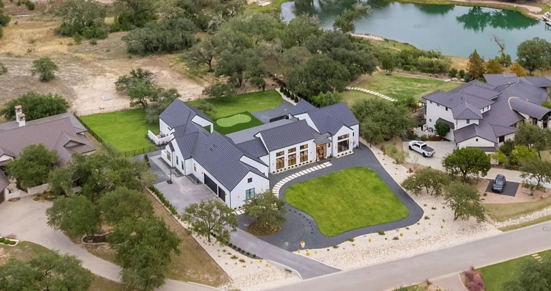 White farmhouse with a dark metal roof at sunset, behind a live oak and a forecourt of gravel and boulders.