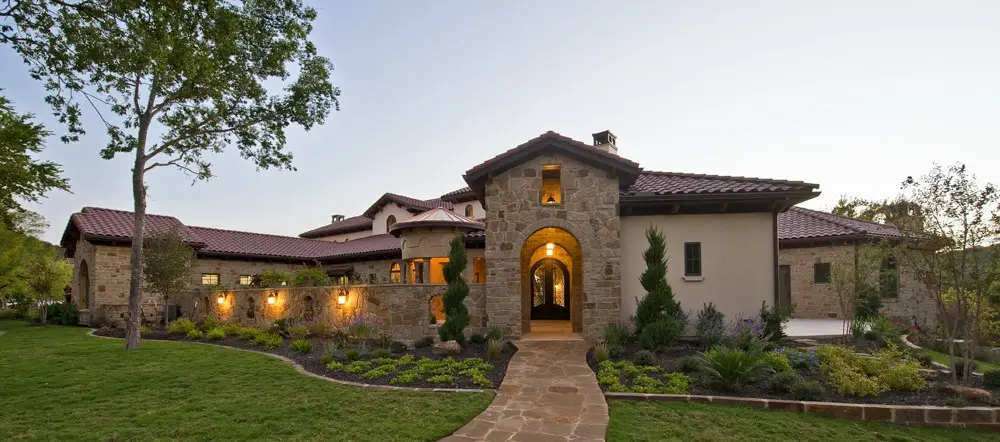 Front elevation at dusk of a stone and stucco home with a tile roof, lit entry arch and a flagstone walk across the lawn.