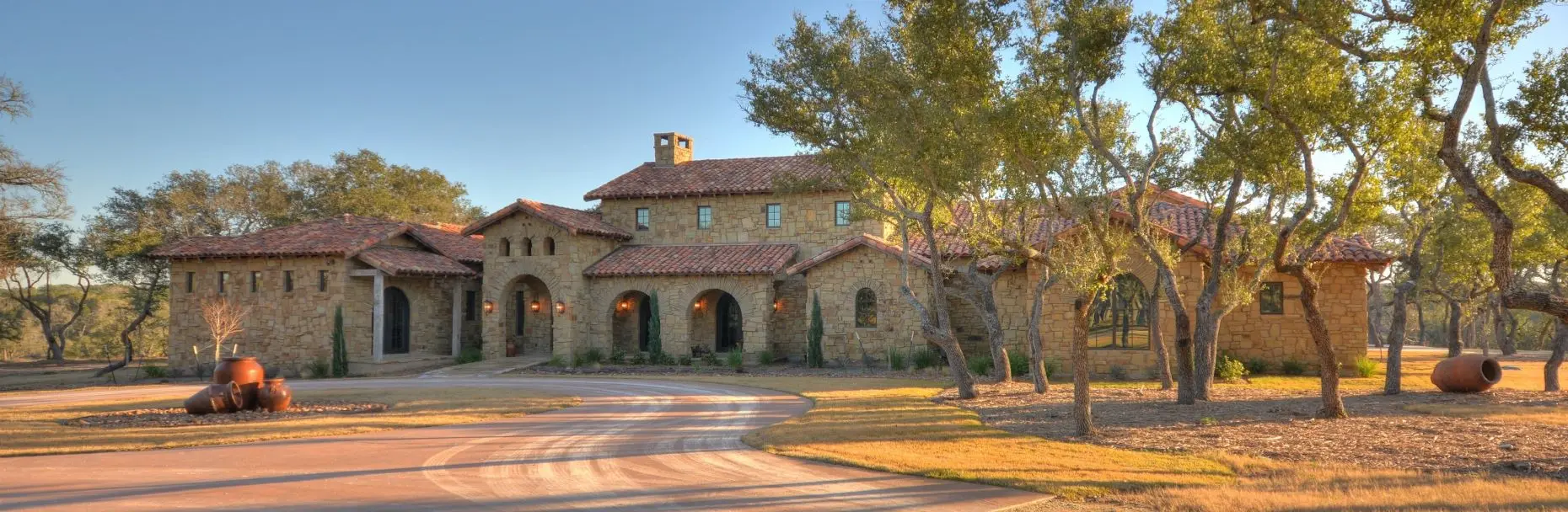 Stone ranch home at dusk with a tile roof and lit arcade, behind a gravel bed and large clay urns in the motor court.
