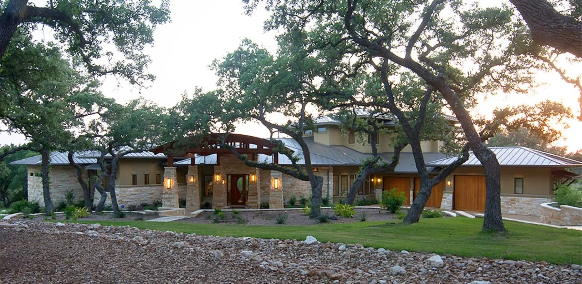 Long stone house with a curved timber entry canopy, seen at dusk through the trunks of live oaks over a gravel bed.