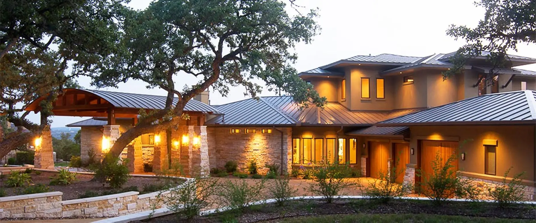 Long stone home with a curved timber entry canopy, seen at dusk through the trunks of live oaks over a gravel bed.