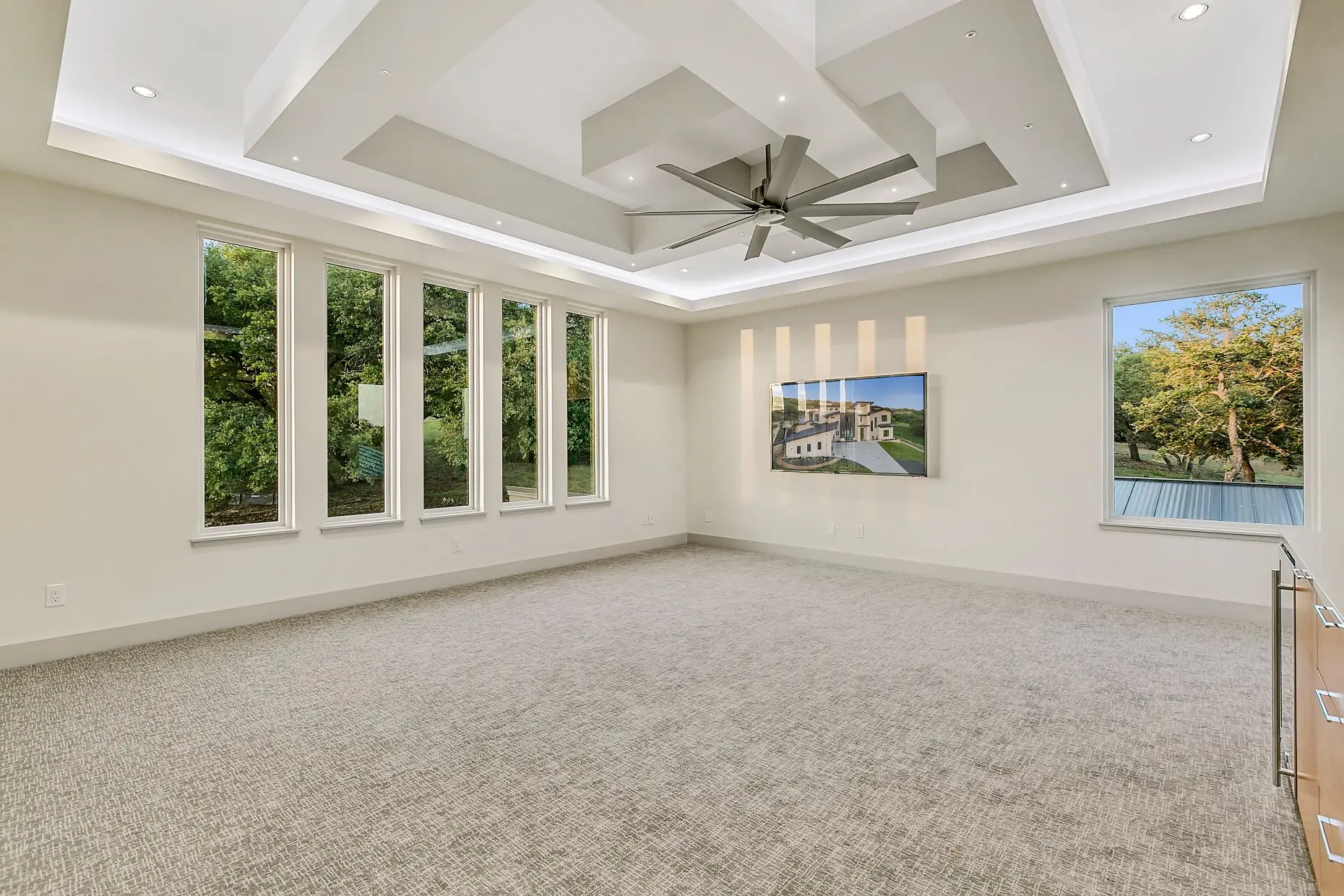 An empty carpeted room with a run of tall windows and a stepped tray ceiling, before any furniture is in it.