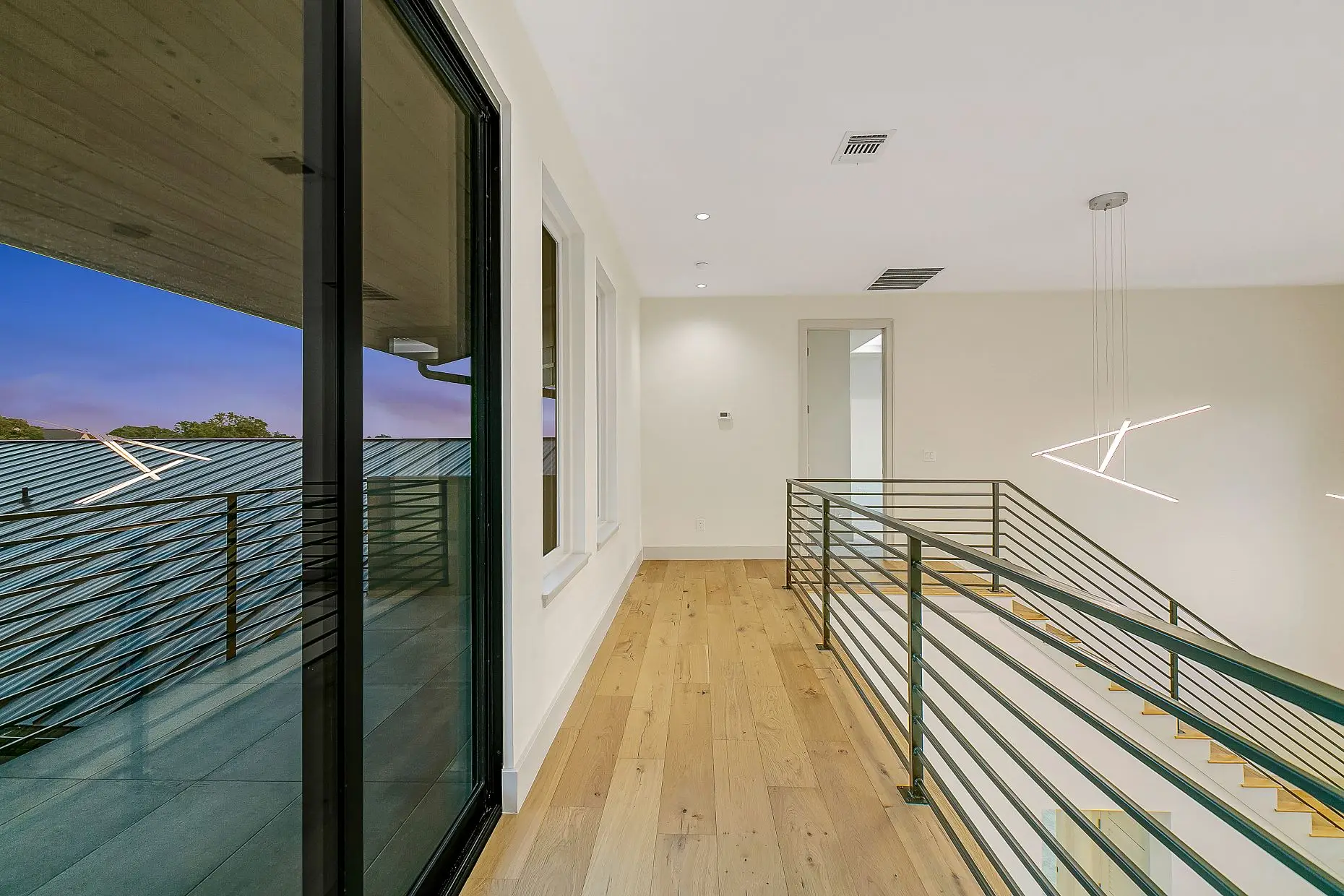 Upper landing with pale oak flooring and a slender metal rail, opening through a glass wall onto a balcony.