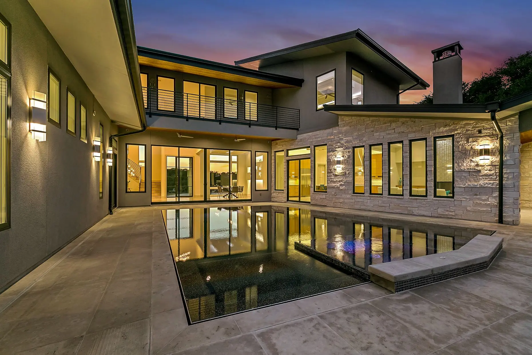 Courtyard pool at dusk reflecting a lit two-storey modern house in dark stucco and stone with deep overhangs.