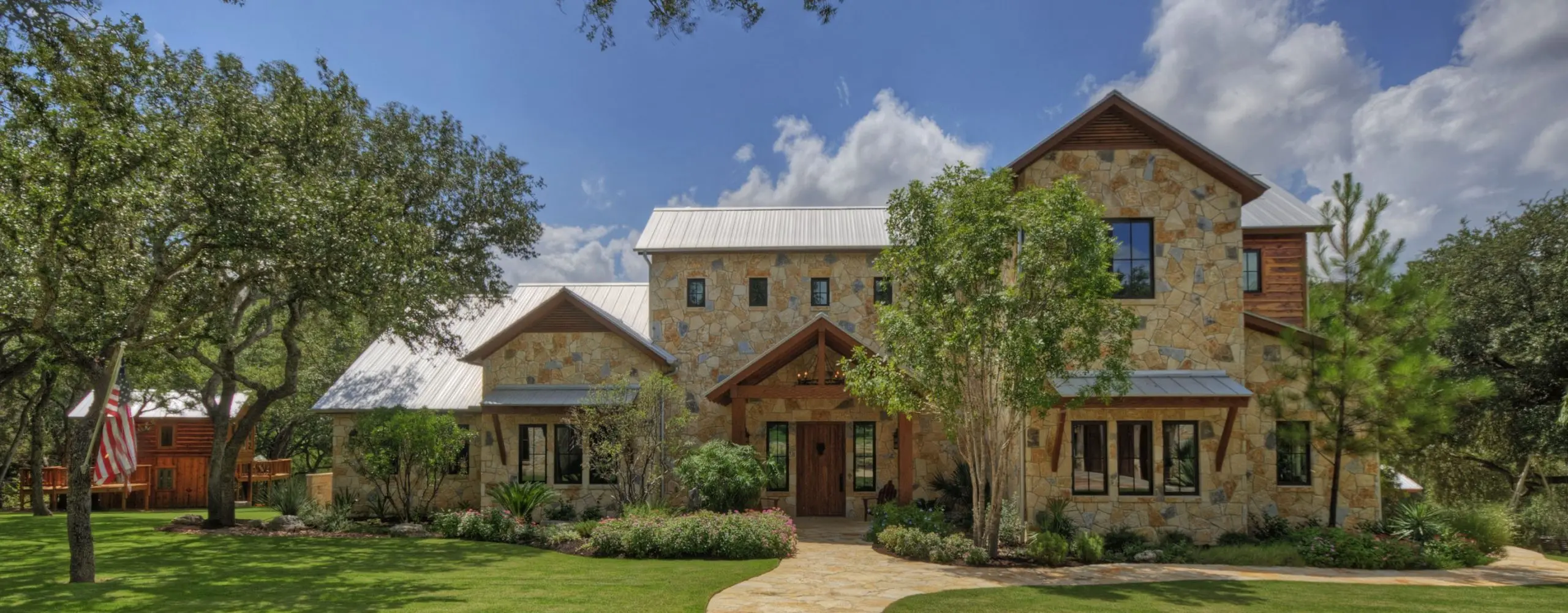 Two-storey stone and timber house with a standing seam metal roof, framed by oaks above a clipped lawn.