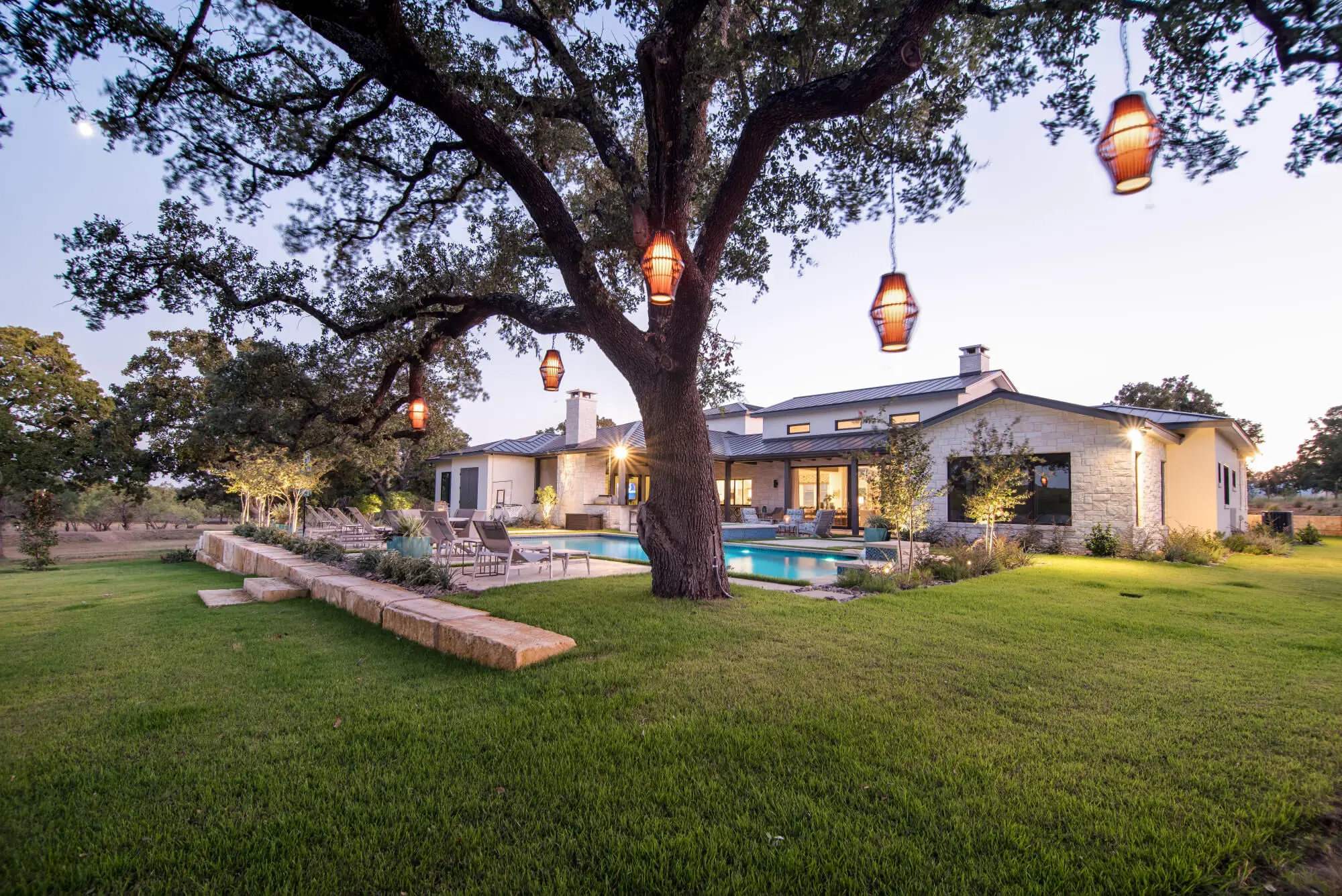 Lit house seen at twilight across a lawn, under a spreading live oak hung with glass pendants.