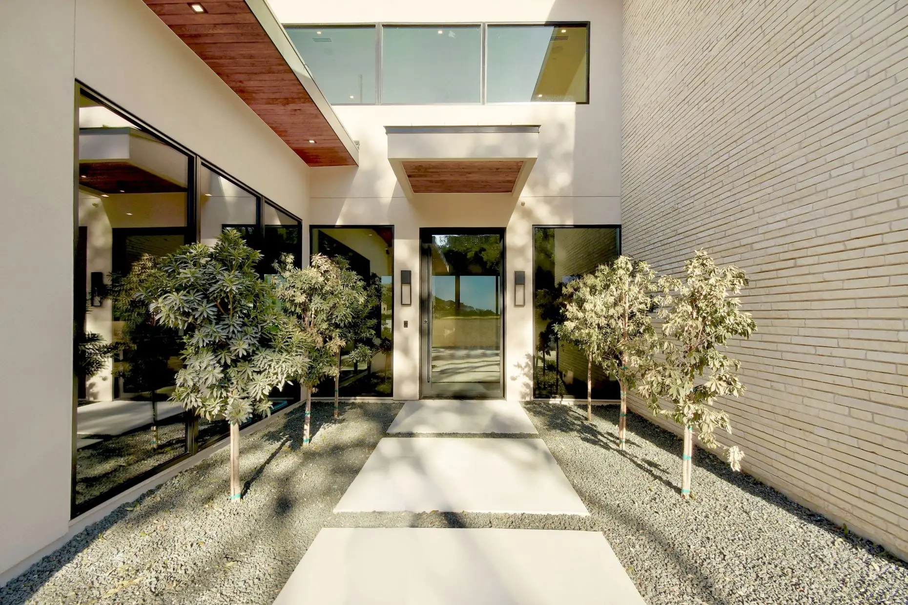 Entry courtyard between a brick wall and a glazed wing, with two slender trees in a gravel bed and a run of pale stone pavers leading to the front door.