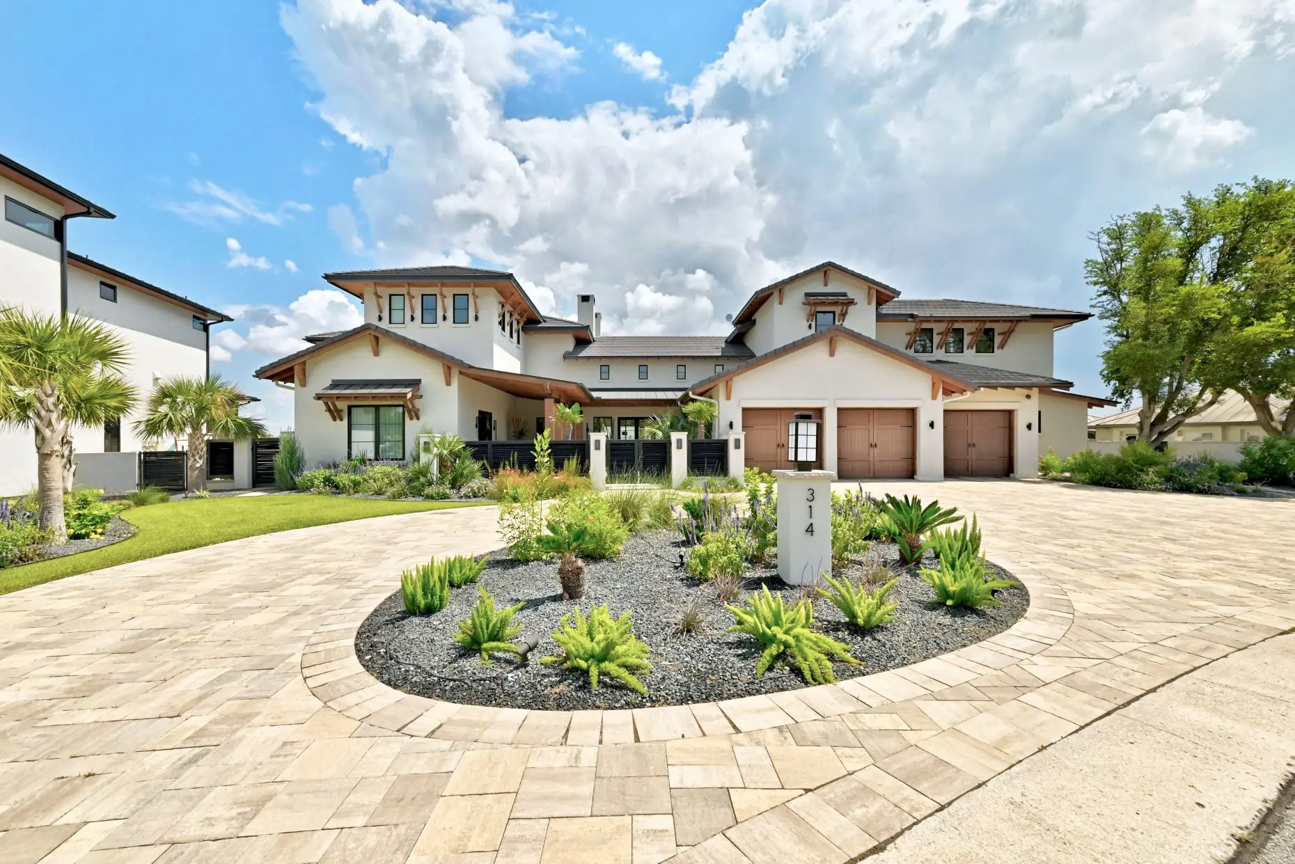 Front elevation behind a circular planted motor court, with a broad stone and stucco facade and palms to one side.