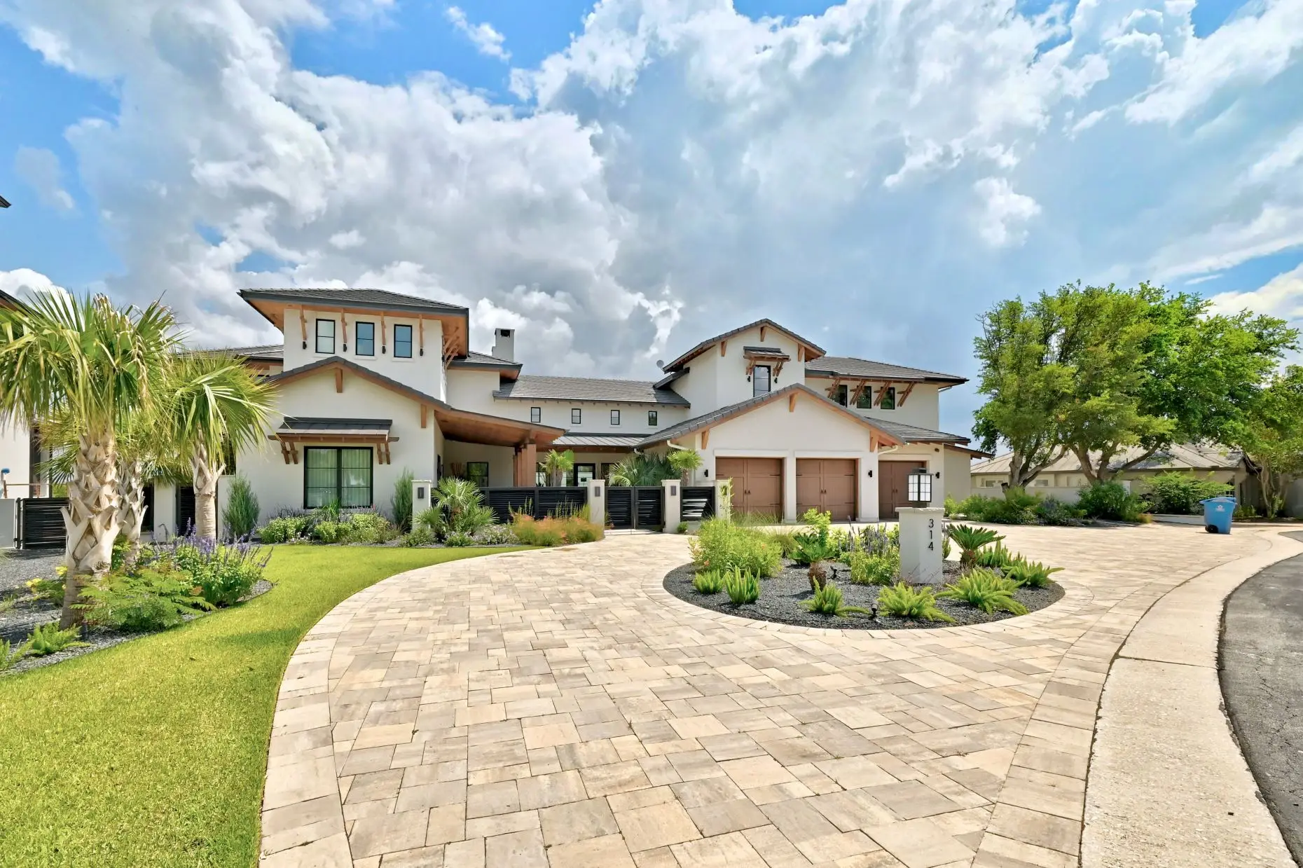 Front elevation behind a circular planted motor court, with a broad stone and stucco facade and palms to one side.