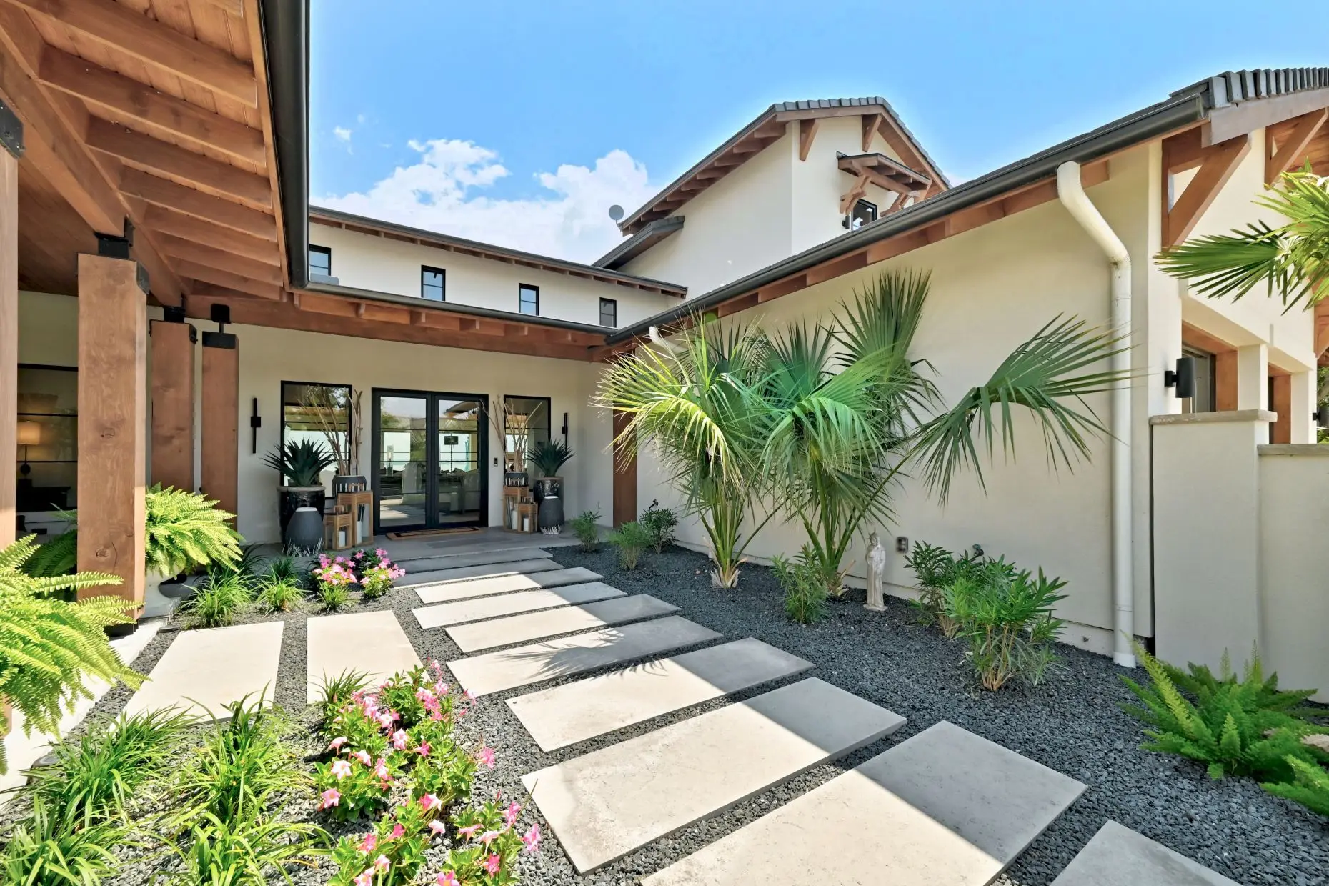 Front elevation behind a circular planted motor court, with a broad stone and stucco facade and palms to one side.