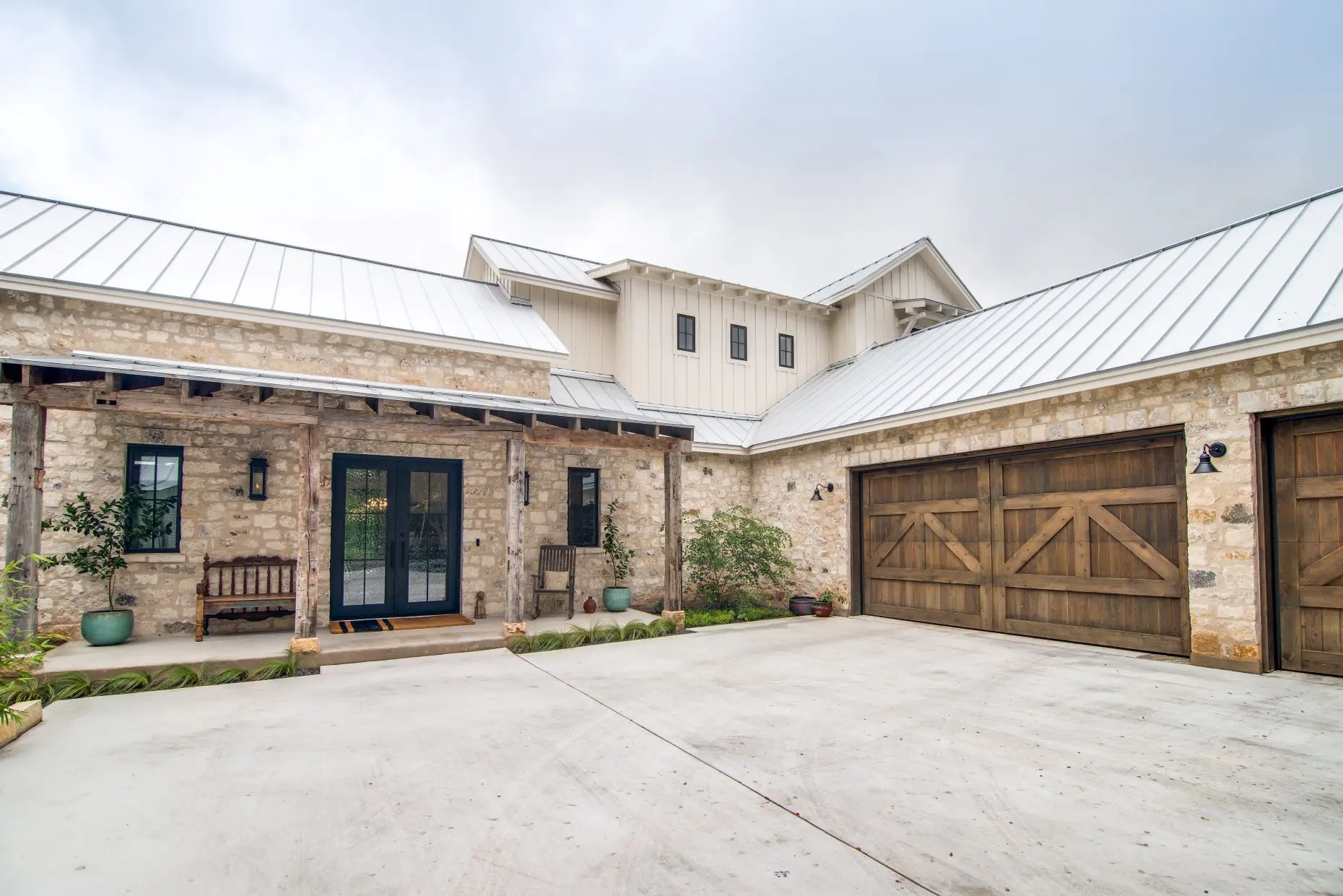 Limestone entry courtyard with a timber-framed porch, black double doors and reclaimed wood garage doors under a standing seam roof.