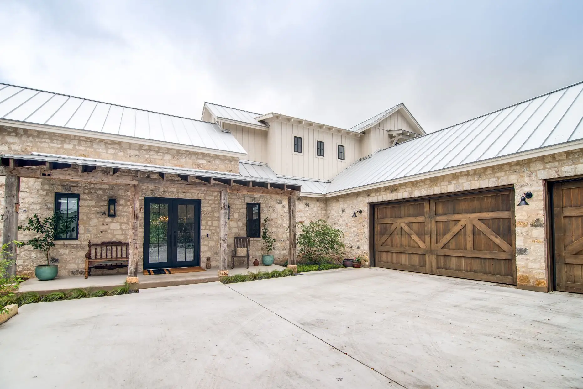 Limestone entry courtyard with a timber-framed porch, black double doors and reclaimed wood garage doors under a standing seam roof.