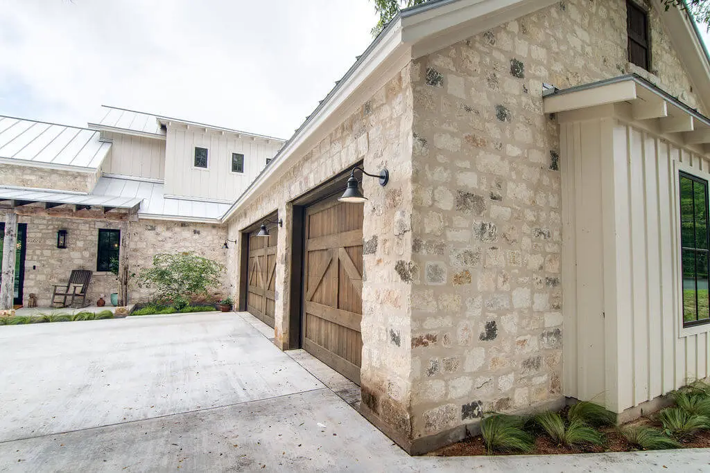 Limestone entry courtyard with a timber-framed porch, black double doors and reclaimed wood garage doors under a standing seam roof.