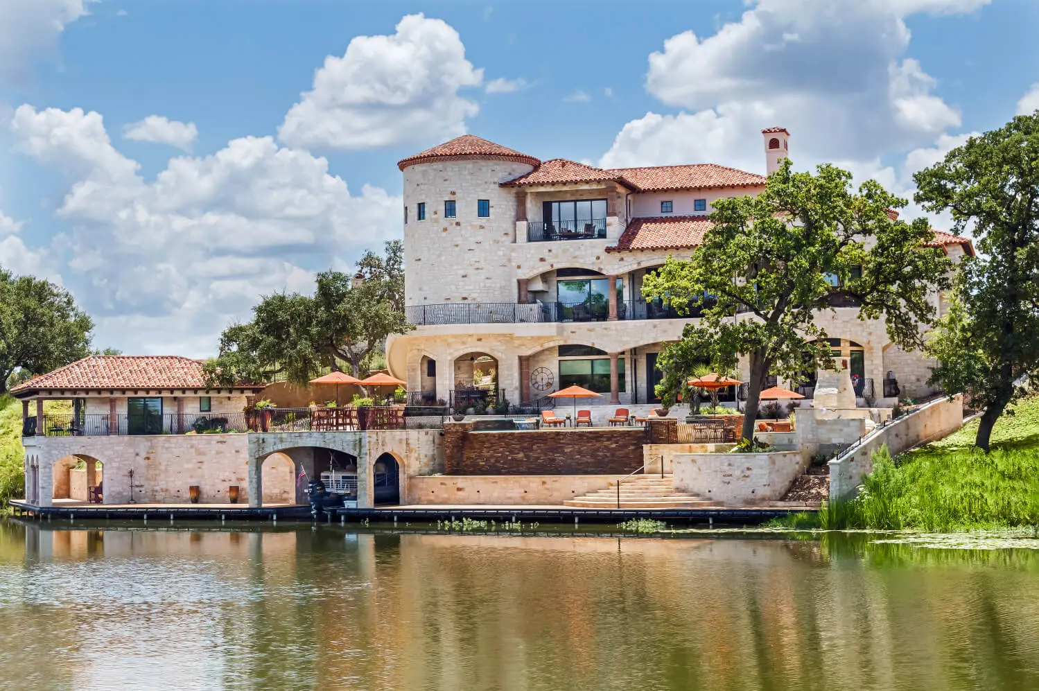 Tuscan-style villa with tile roofs and a stone terrace, seen across still water from the far bank.
