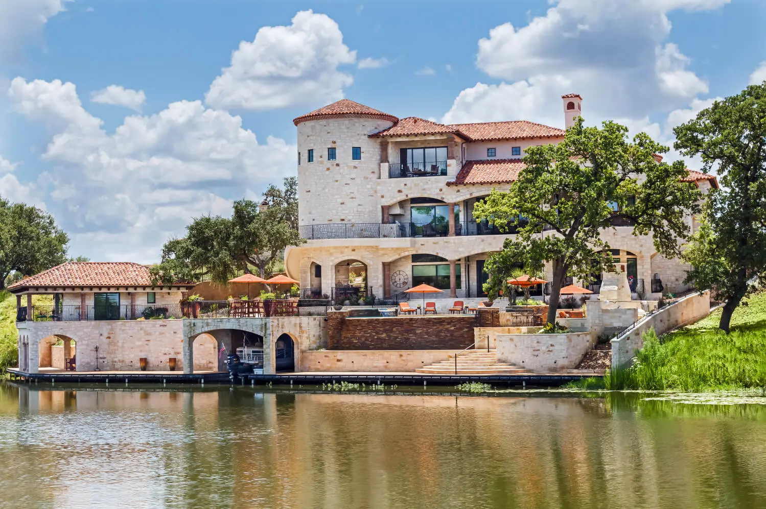 Tuscan-style villa with tile roofs and a stone terrace, seen across still water from the far bank.