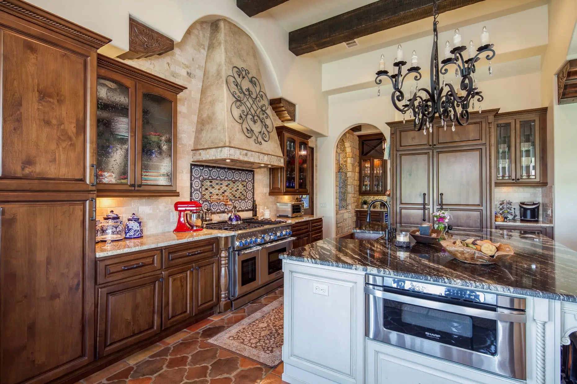 Great room under a beamed ceiling, with a carved stone fireplace, arched openings and a wall of built-in cabinets.