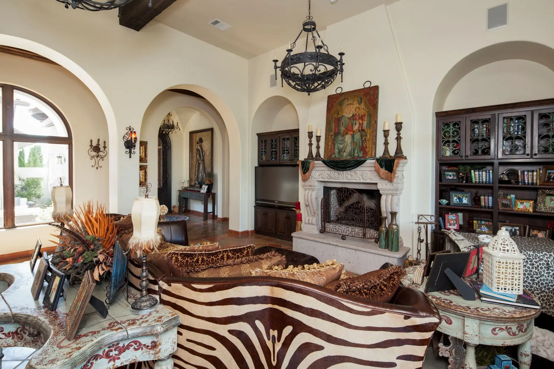 Great room under a beamed ceiling, with a carved stone fireplace, arched openings and a wall of built-in cabinets.