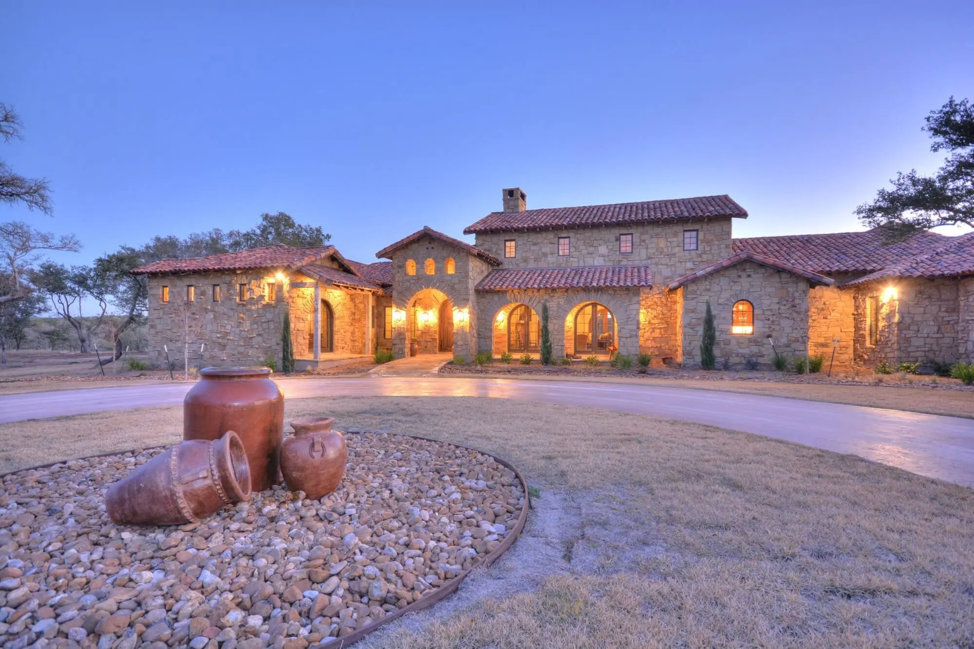 Stone ranch house at dusk with a tile roof and lit arcade, behind a gravel bed and large clay urns in the motor court.