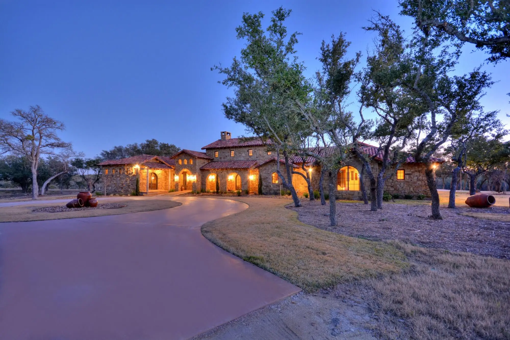 Stone and timber ranch home lit from within at dusk, seen across a curving drive under scattered live oaks.