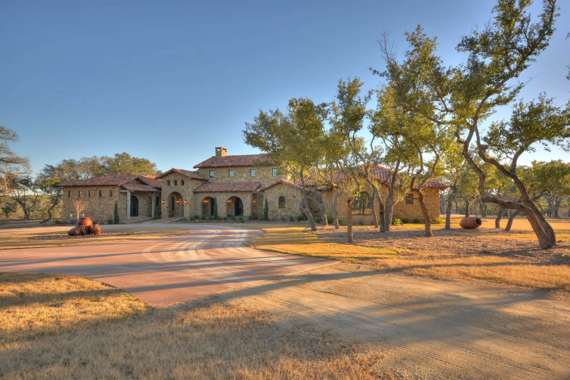 Stone ranch home at dusk with a tile roof and lit arcade, behind a gravel bed and large clay urns in the motor court.