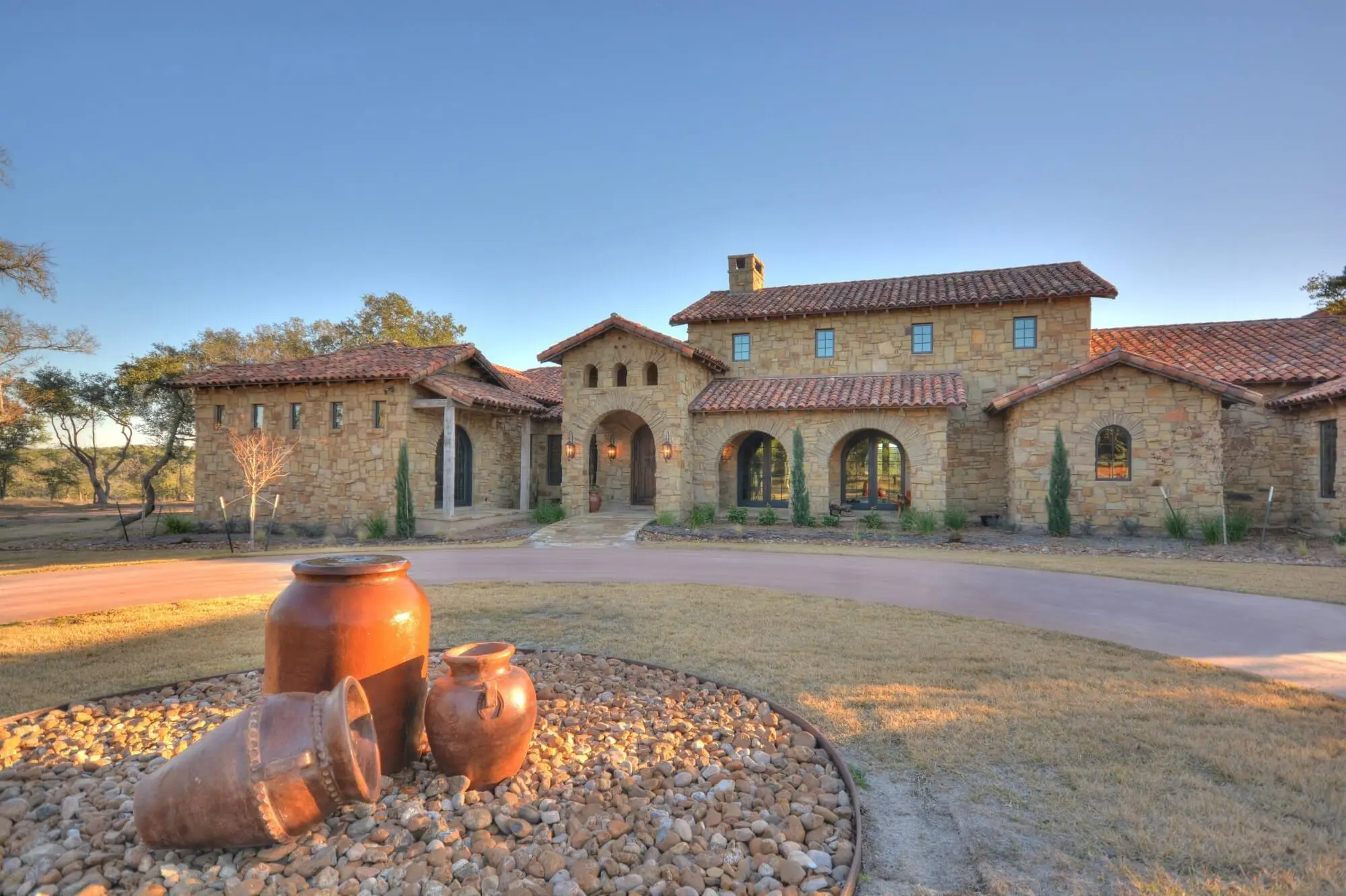 Stone ranch home at dusk with a tile roof and lit arcade, behind a gravel bed and large clay urns in the motor court.