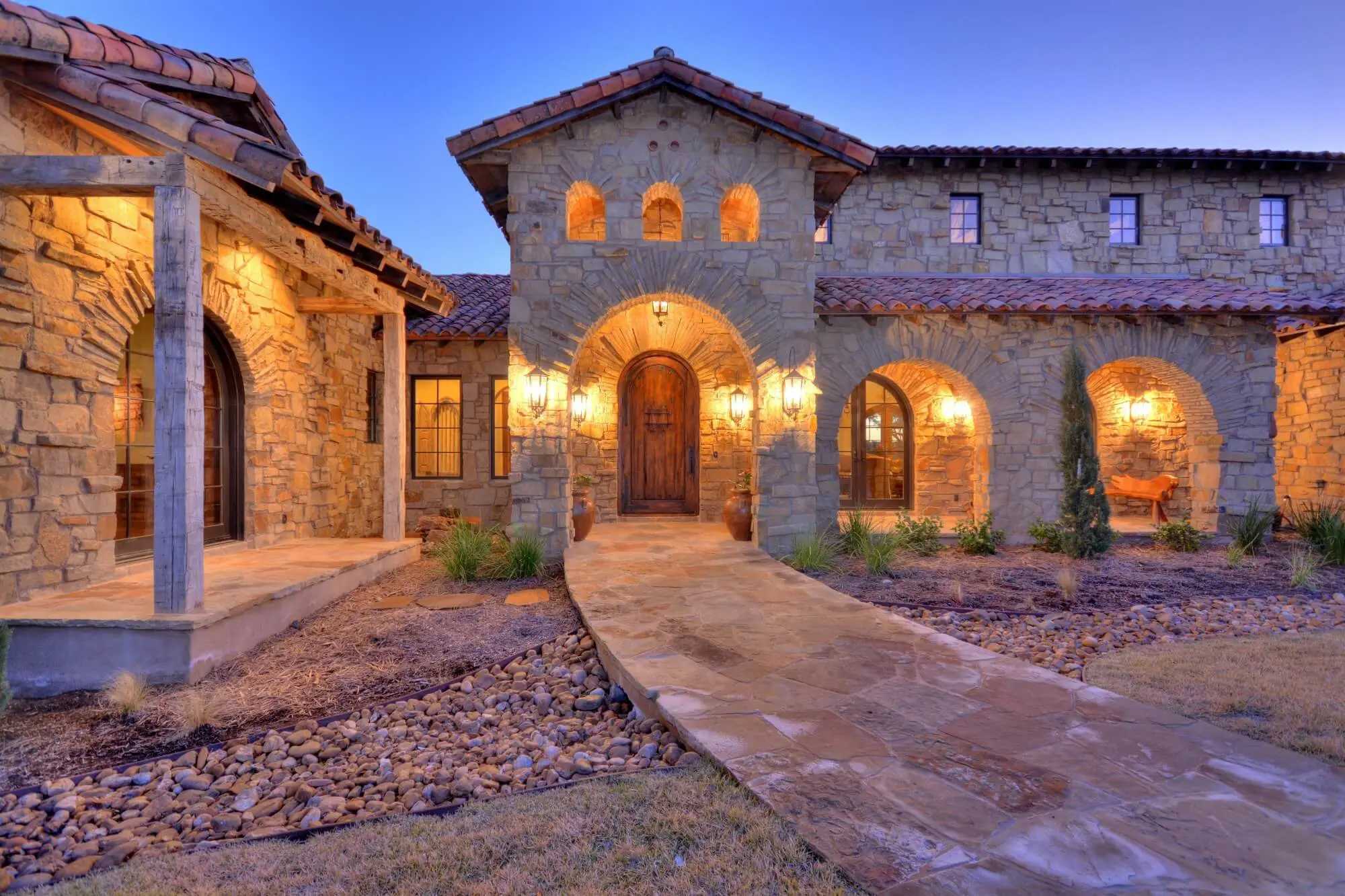 Stone ranch home at dusk with a tile roof and lit arcade, behind a gravel bed and large clay urns in the motor court.