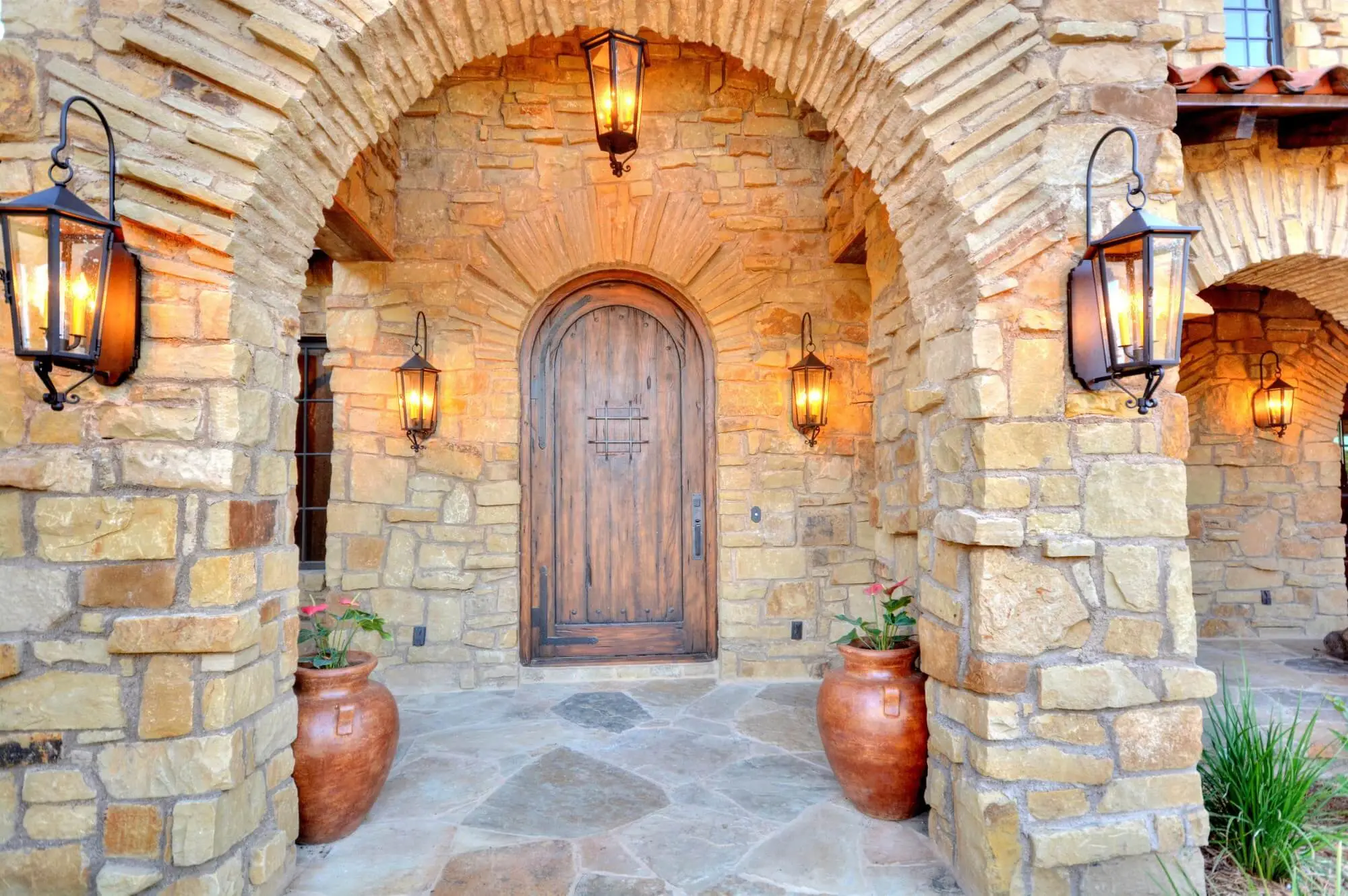 Stone ranch home at dusk with a tile roof and lit arcade, behind a gravel bed and large clay urns in the motor court.