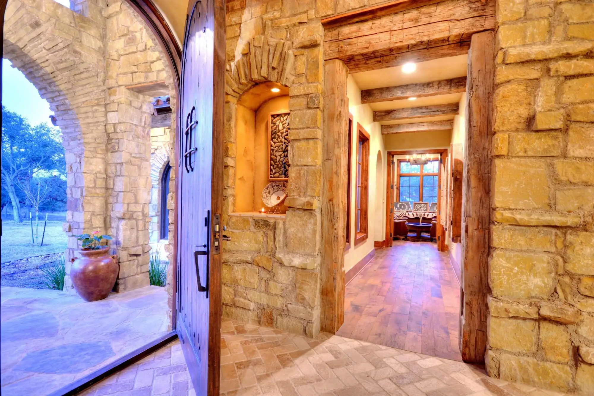 Stone ranch home at dusk with a tile roof and lit arcade, behind a gravel bed and large clay urns in the motor court.