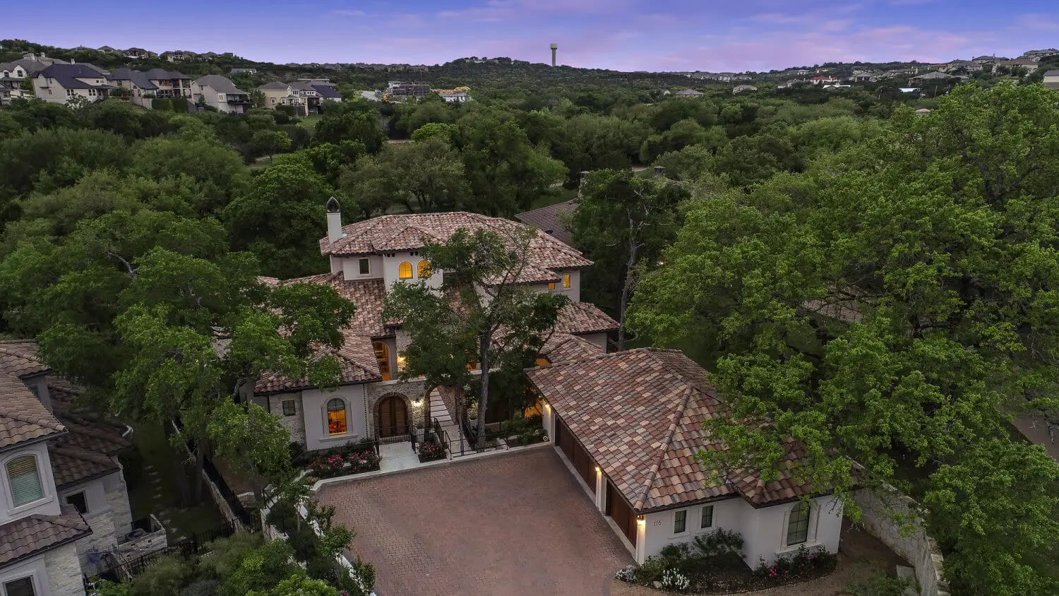 Aerial view at dusk of a tile-roofed home wrapped around a lit pool and spa, surrounded by oaks.