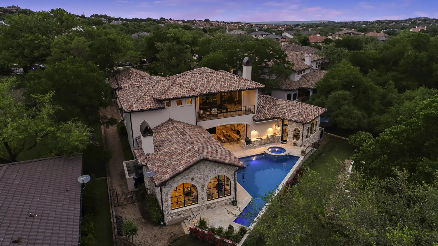 Aerial view at dusk of a tile-roofed home wrapped around a lit pool and spa, surrounded by oaks.