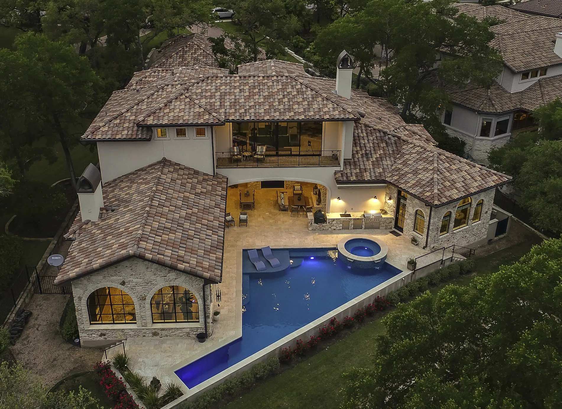 Aerial view at dusk of a tile-roofed house wrapped around a lit pool and spa, surrounded by oaks.