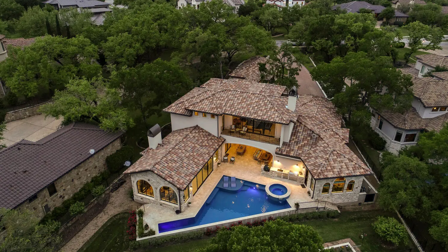 Aerial view at dusk of a tile-roofed home wrapped around a lit pool and spa, surrounded by oaks.