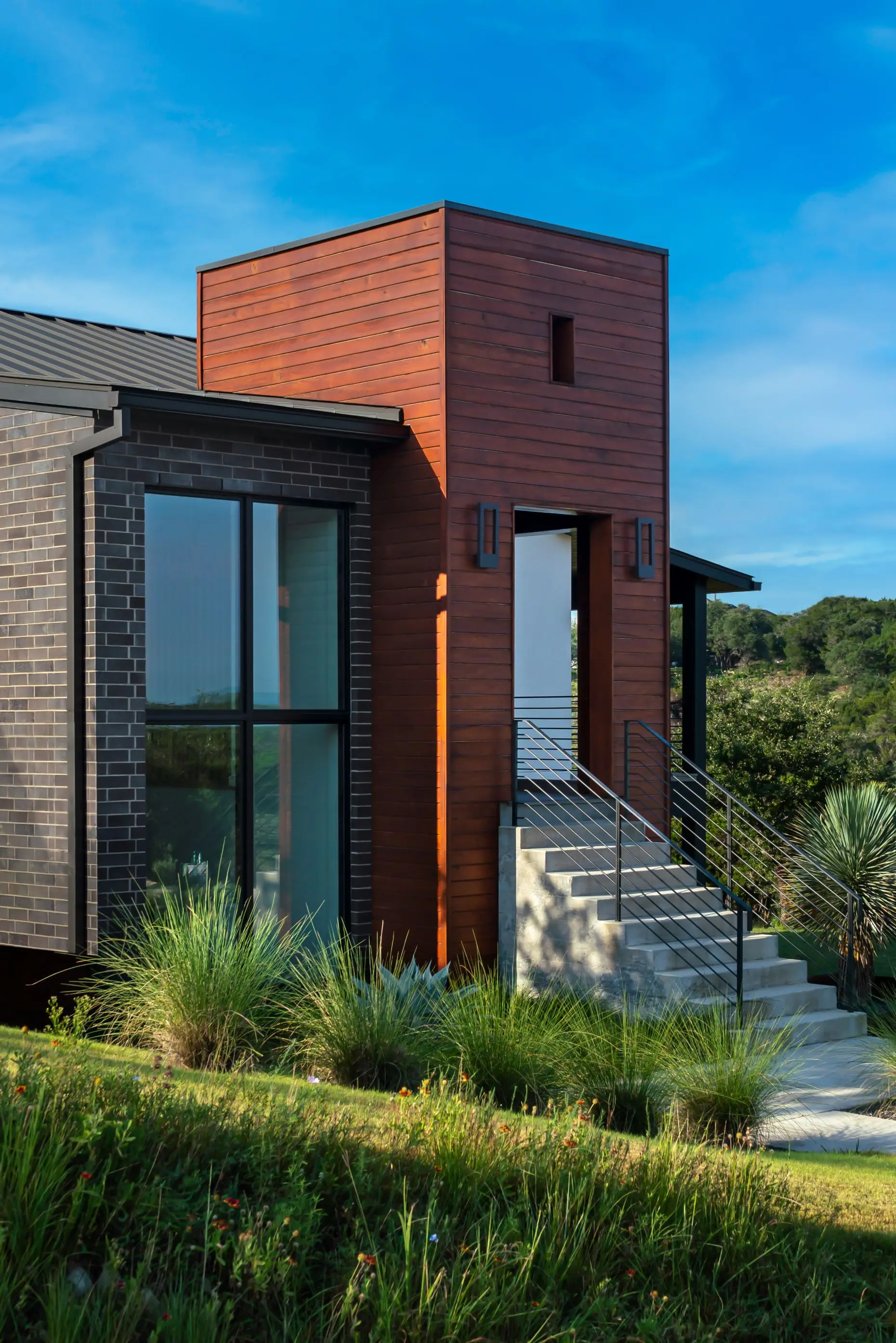 Street elevation at dusk of a low modern home with dark cladding and a metal roof, set above a sloping drive.