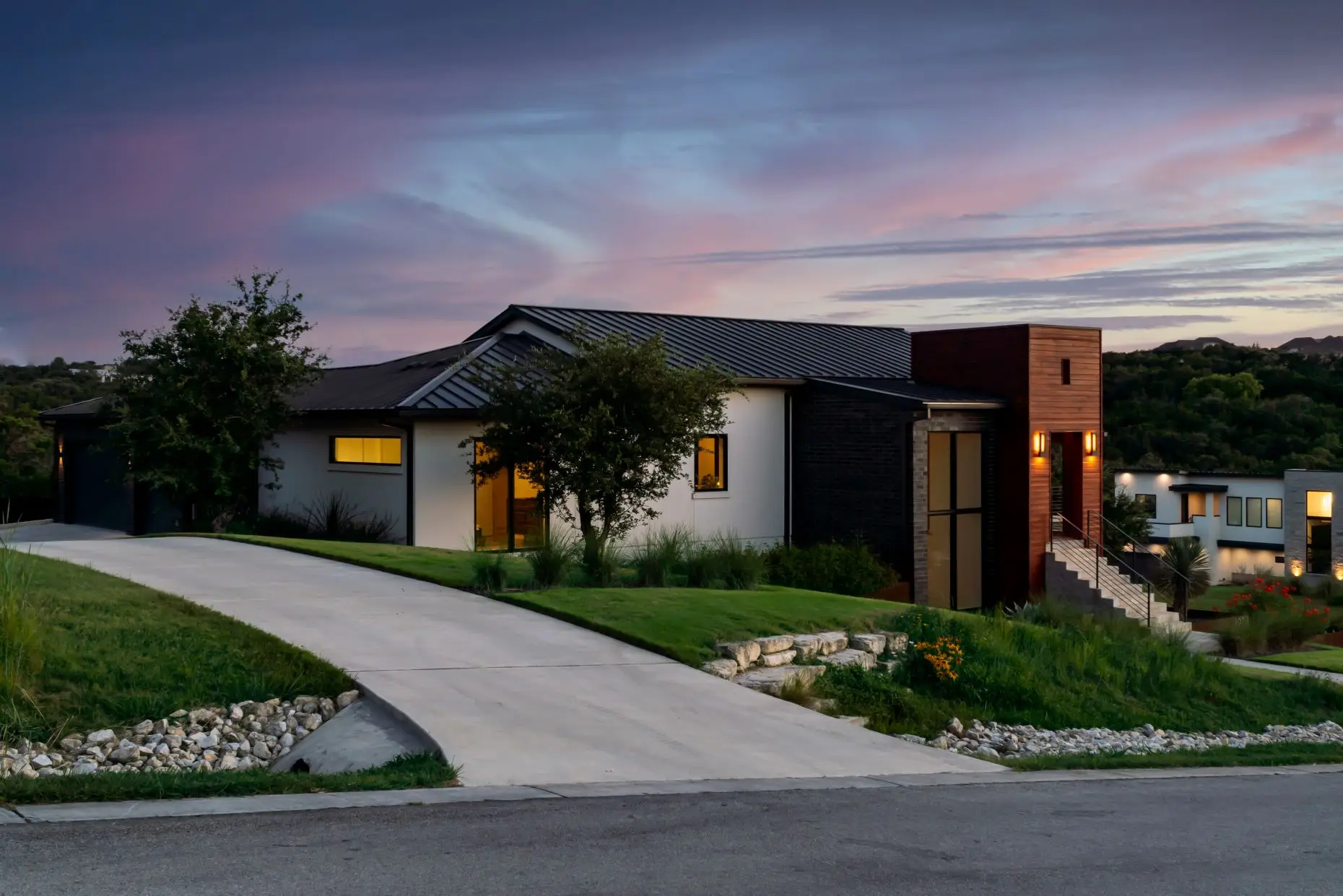 Street elevation at dusk of a low modern house with dark cladding and a metal roof, set above a sloping drive.