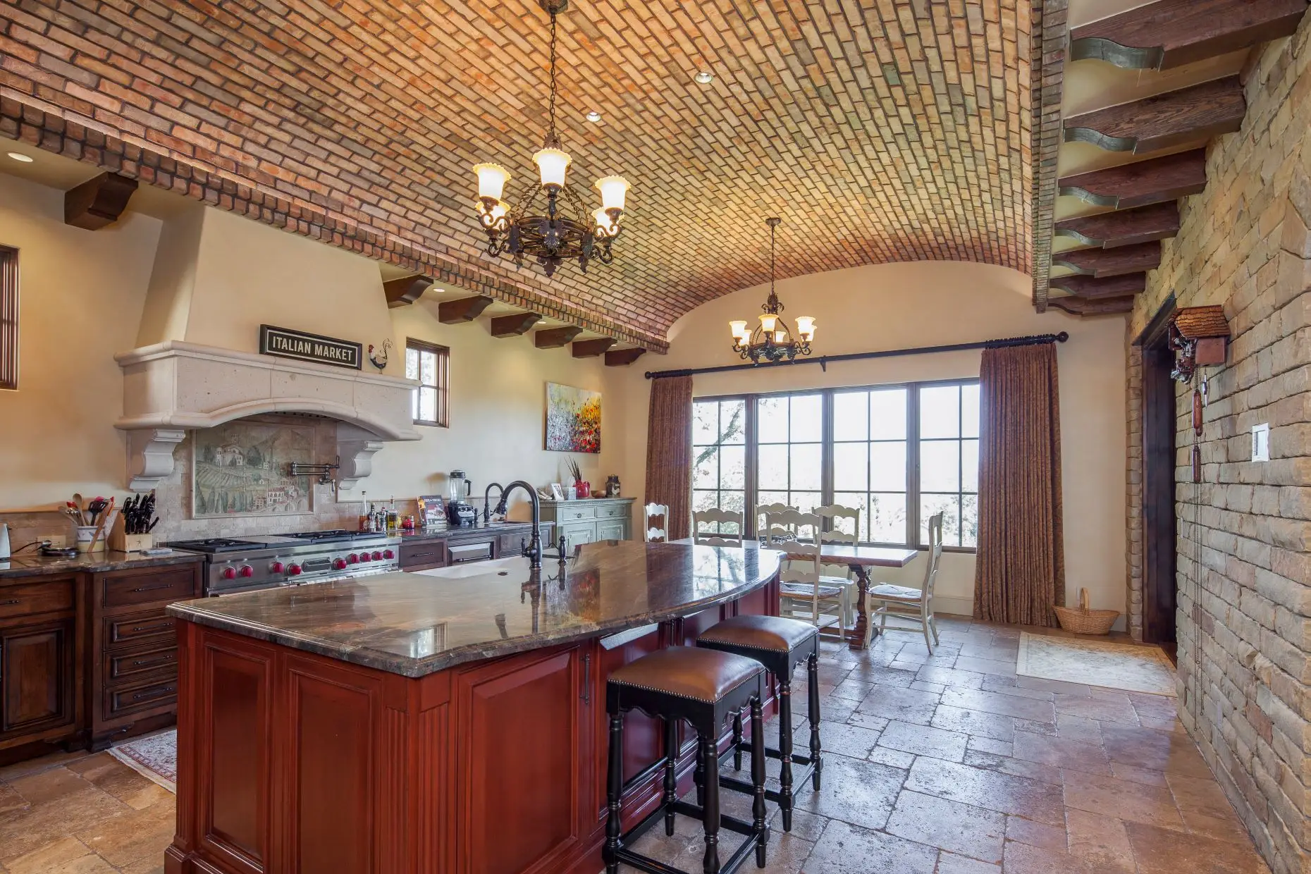 Kitchen under a brick barrel-vaulted ceiling, with a stone range hood, a long island with stools and a window onto open country.