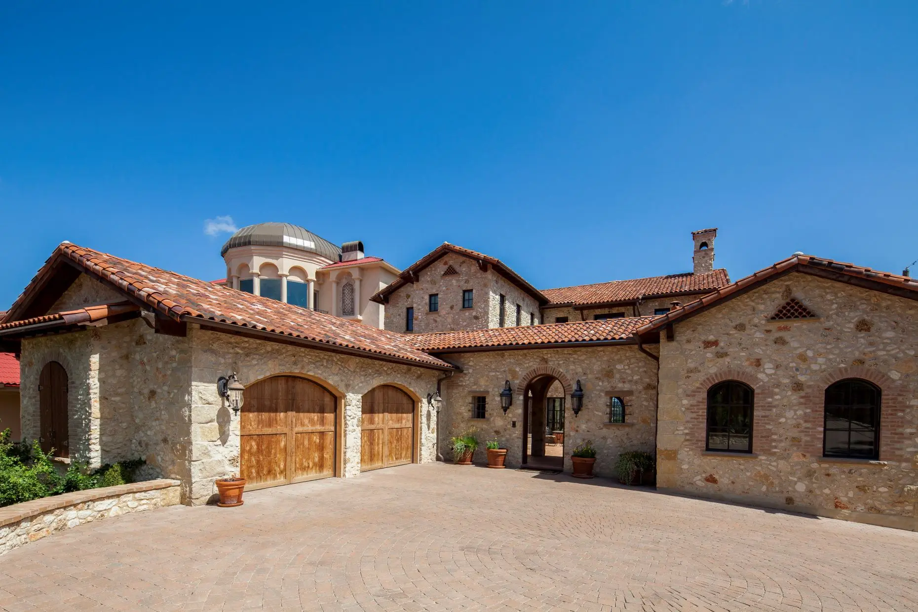 Stone house with a tile roof and arched timber garage doors, around a paved motor court.