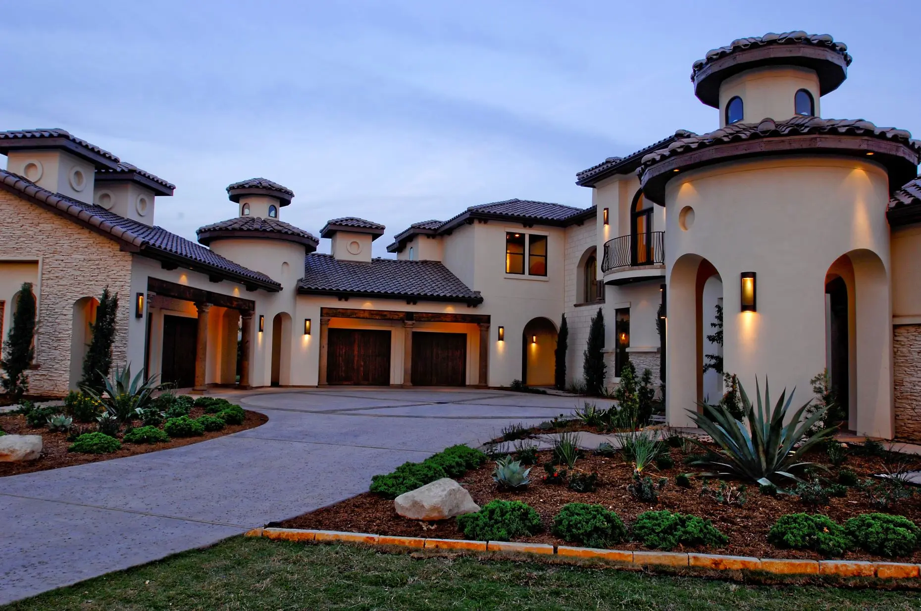 Front elevation at dusk of a Mediterranean house with a stucco turret, tile roof and lit arched entry above a curved drive.