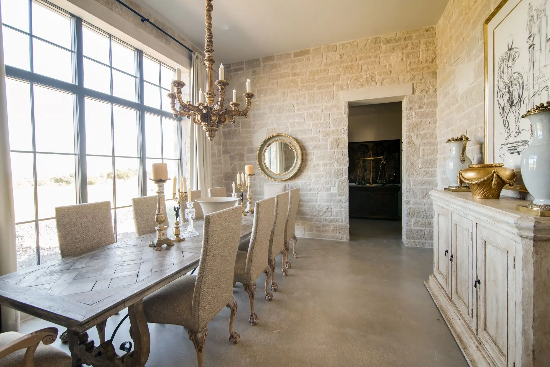 Dining room with a long timber table and high-backed chairs, set against a coursed limestone wall under an iron chandelier, with tall windows along one side.