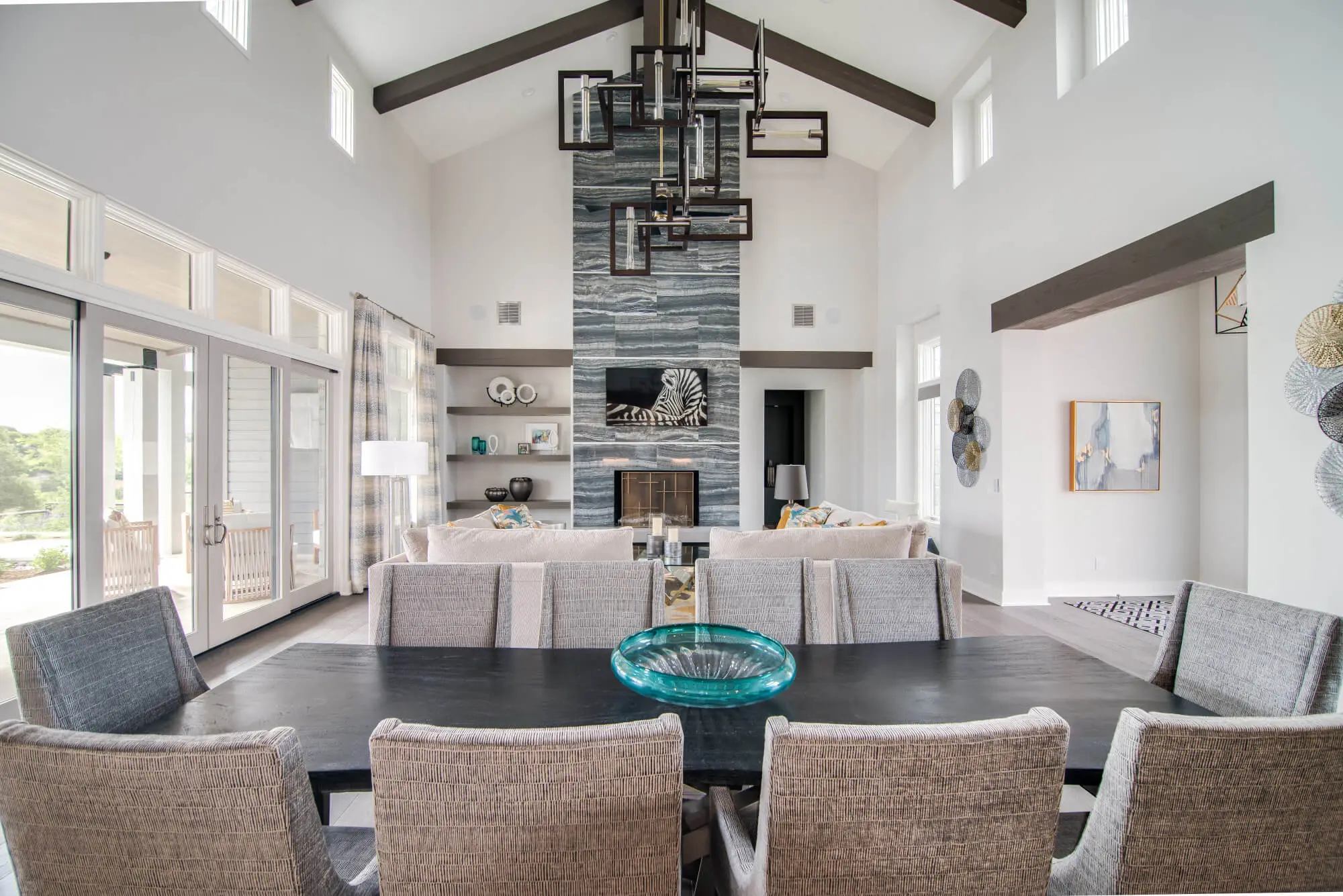 A long dining table with upholstered chairs under a vaulted ceiling with dark beams, beside a full-height stone fireplace.