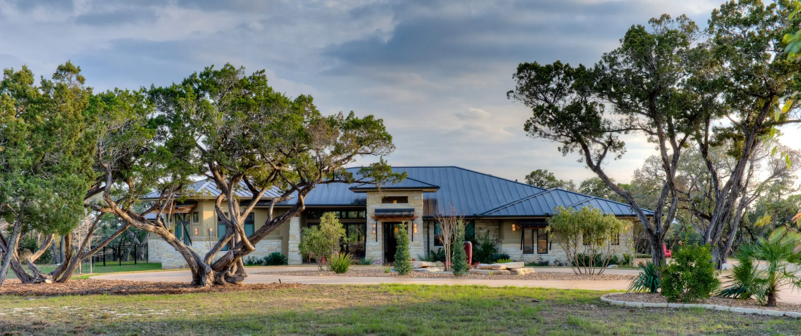 Long low house with a metal roof and stone base, seen across open ground between two oaks.