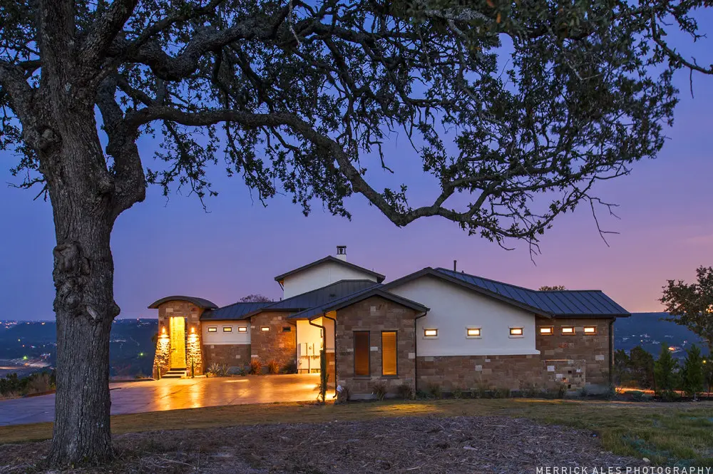 Front elevation at dusk of a contemporary stone and stucco home with a metal roof and lit garage bays across a wide drive.