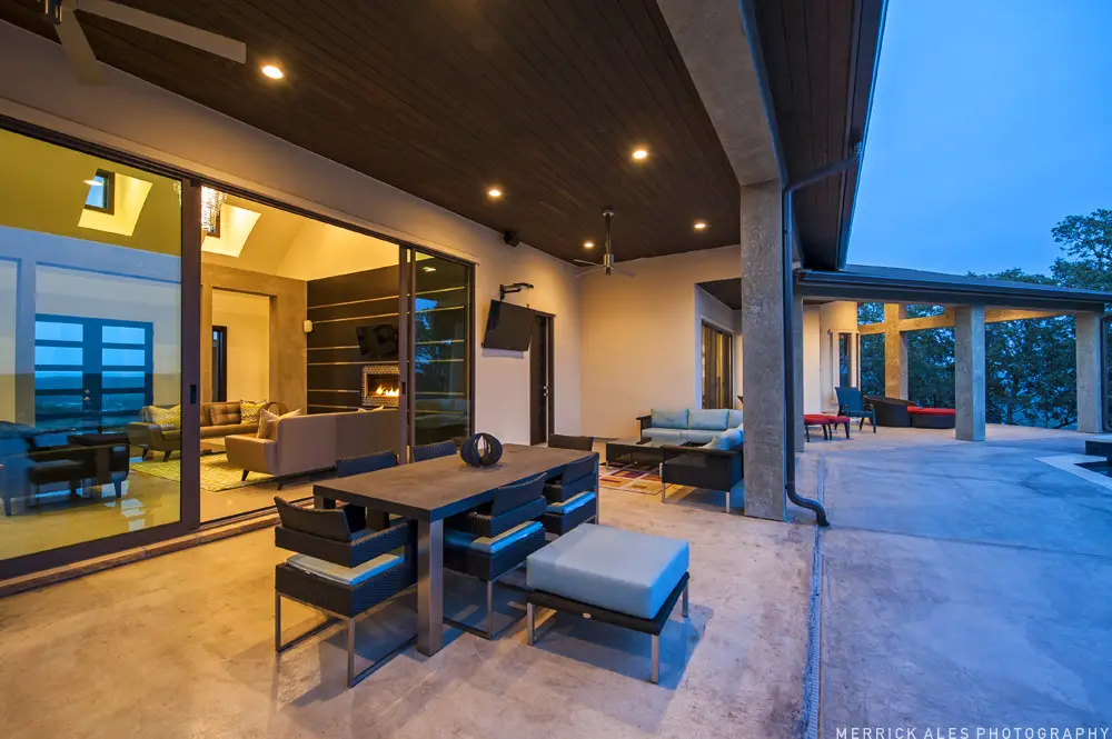 Front elevation at dusk of a contemporary stone and stucco home with a metal roof and lit garage bays across a wide drive.