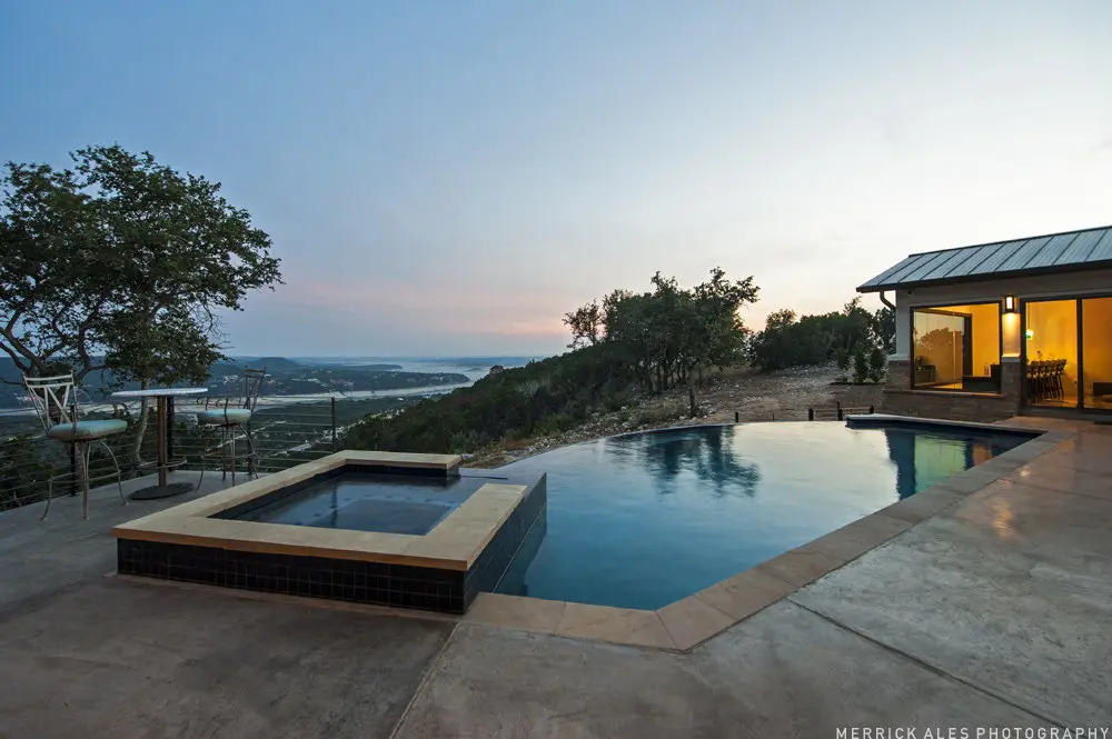 Front elevation at dusk of a contemporary stone and stucco home with a metal roof and lit garage bays across a wide drive.