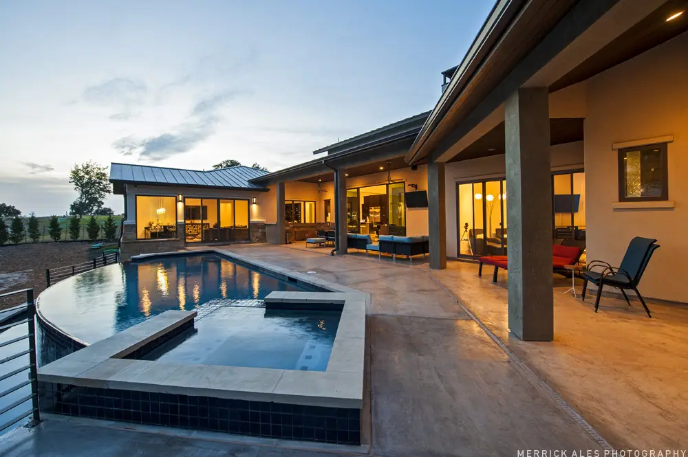 Front elevation at dusk of a contemporary stone and stucco home with a metal roof and lit garage bays across a wide drive.