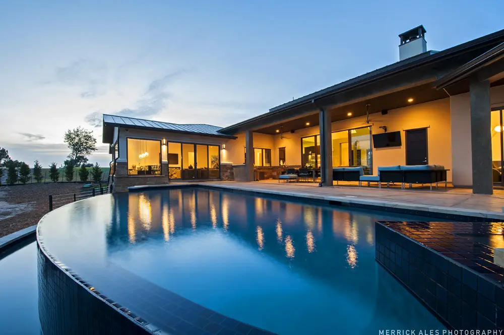 Front elevation at dusk of a contemporary stone and stucco home with a metal roof and lit garage bays across a wide drive.