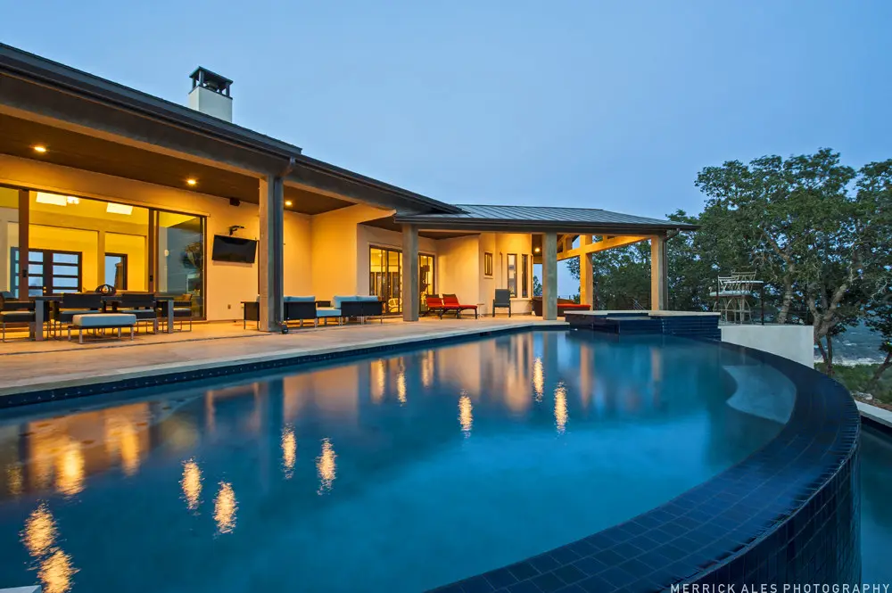 Front elevation at dusk of a contemporary stone and stucco home with a metal roof and lit garage bays across a wide drive.
